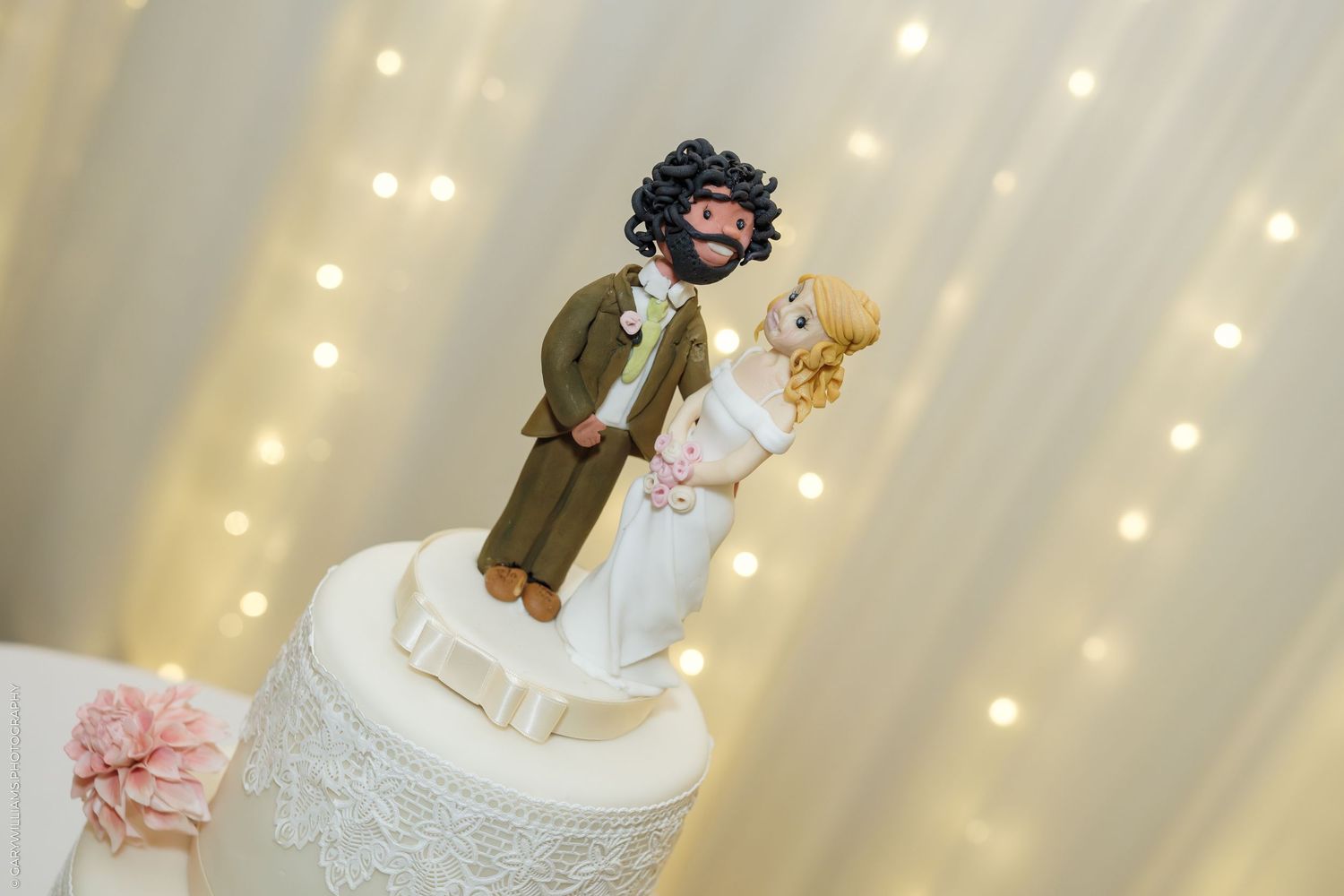 Custom bride and groom wedding cake toppers on a white lace cake with fairy lights backdrop.