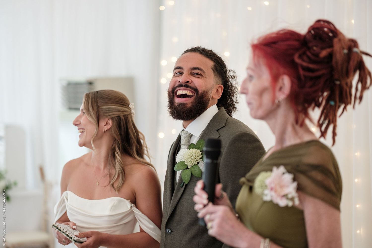 Groom in gray suit laughing joyfully with bride in white dress and bridesmaid with red dreadlocks at wedding ceremony.