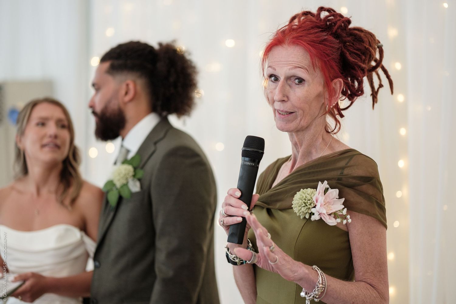 Woman with red dreadlocks giving a speech at a wedding, holding a microphone, wearing an olive green dress.