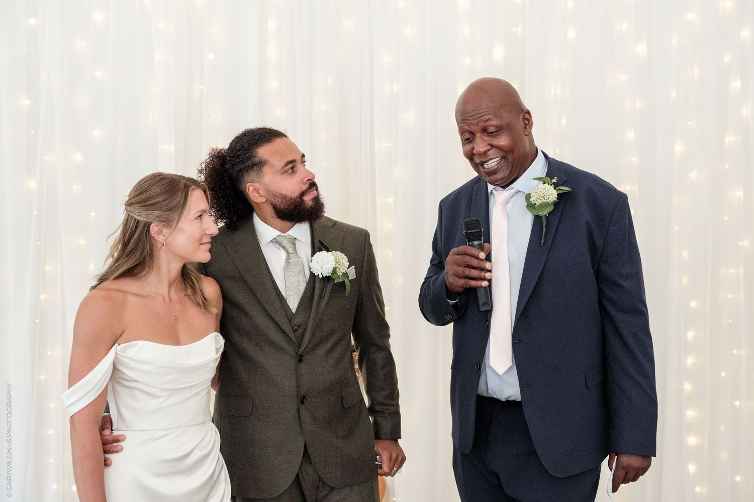 Bride and groom smile with a guest giving a speech at an elegant wedding reception with fairy light backdrop.