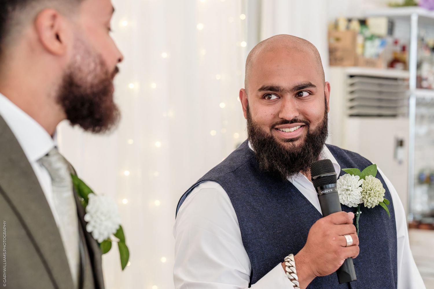 Bald bearded man in vest holds microphone at wedding ceremony, smiling at another man with beard wearing boutonniere.