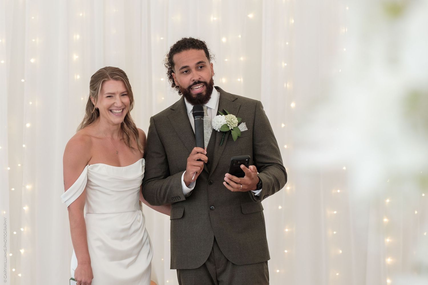 Bride in white off-shoulder gown and groom in olive suit smile during wedding ceremony with fairy lights.