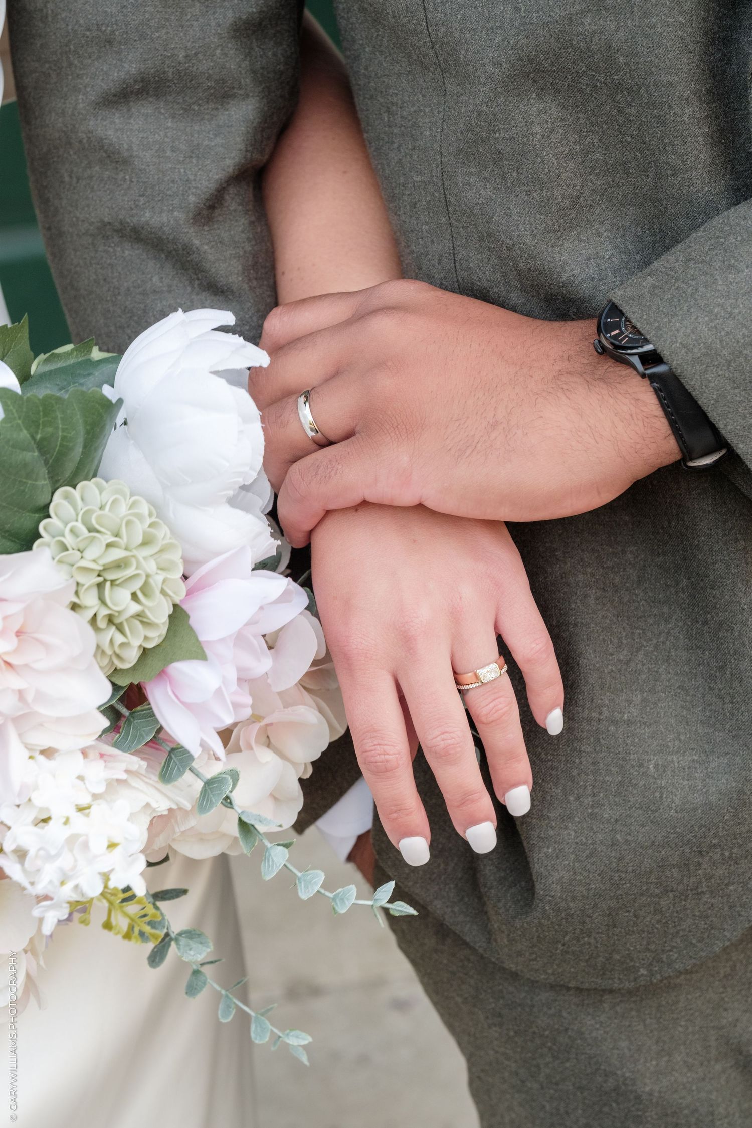 Newlywed couple displaying wedding rings on clasped hands over a bouquet of white and pink flowers.