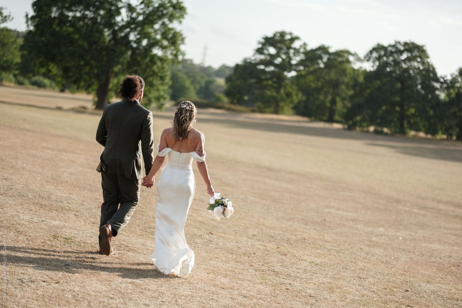 Bride and groom walking hand in hand across a sunlit field, bride holding a white bouquet in an off-shoulder gown.