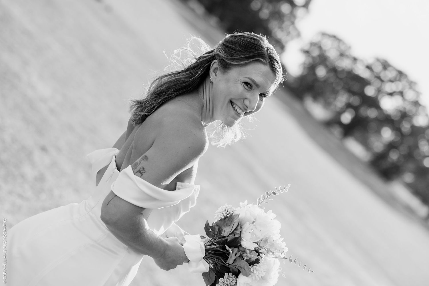 Smiling bride in off-shoulder wedding dress holding bouquet, black and white outdoor portrait.