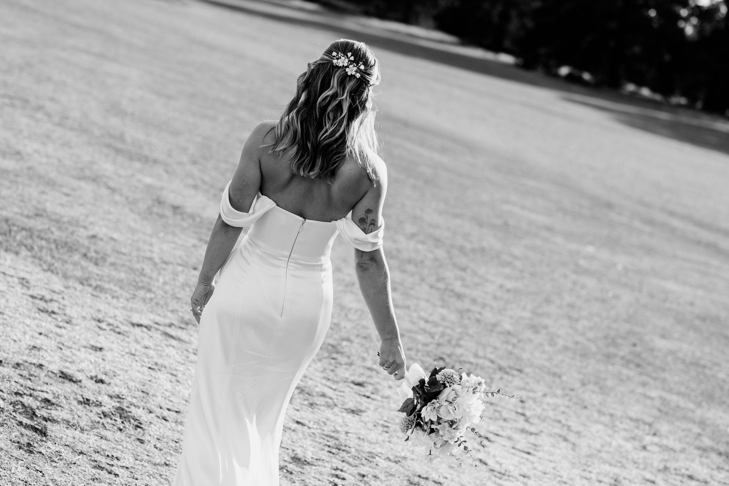 Bride in white off-shoulder gown holding bouquet walks across sunlit field in elegant black and white wedding photo.
