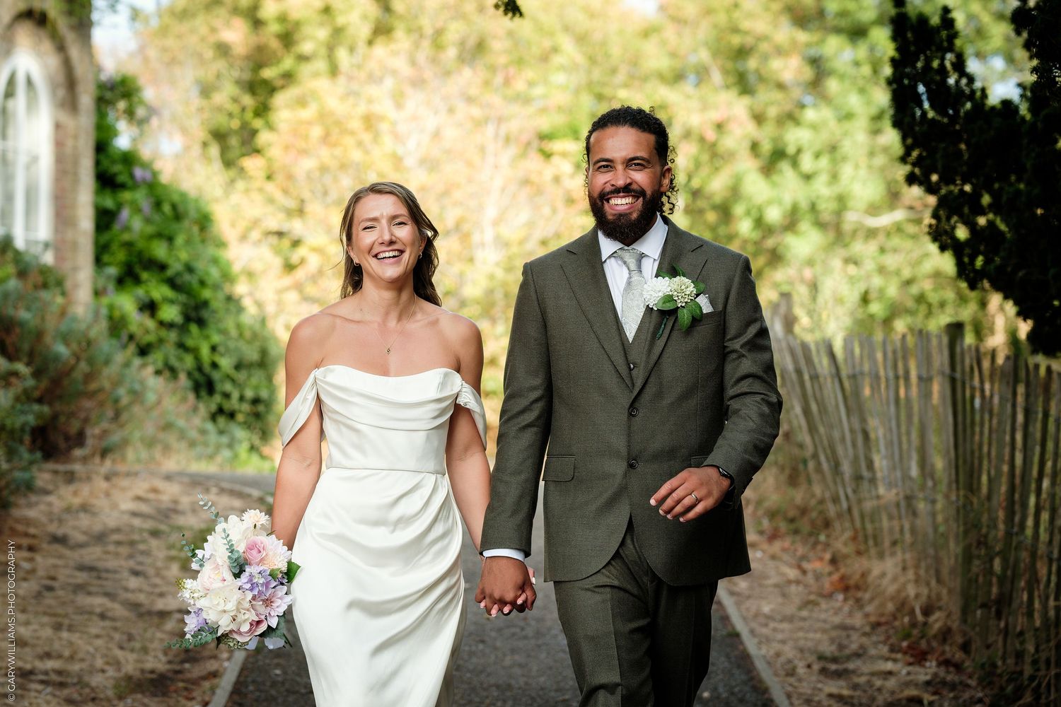 Happy newlyweds walk hand-in-hand, bride in white off-shoulder gown holding bouquet, groom in olive suit, outdoors.