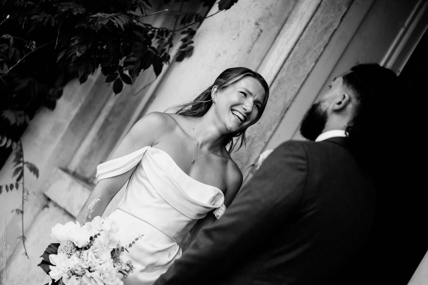 Black and white photo of a laughing bride in strapless gown holding bouquet, facing groom in suit outdoors.