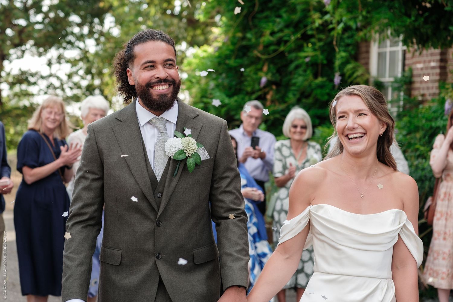Smiling newlywed couple walking hand in hand outdoors, groom in olive suit, bride in white off-shoulder gown, guests applauding.