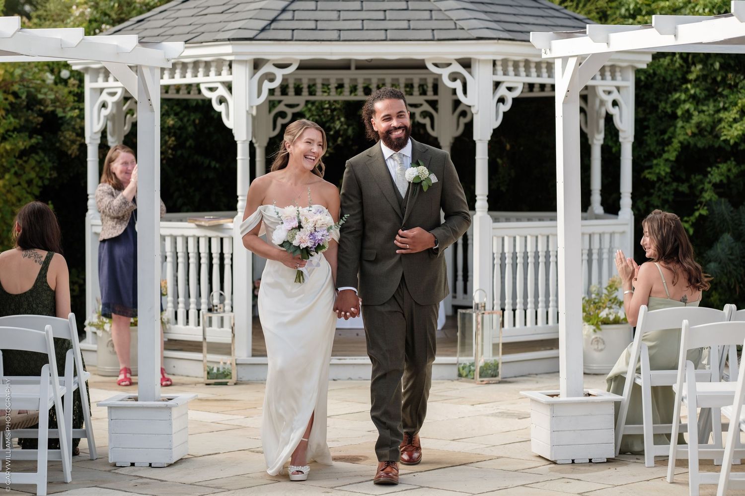 Bride and groom walk hand-in-hand after wedding ceremony at elegant white gazebo outdoor venue.