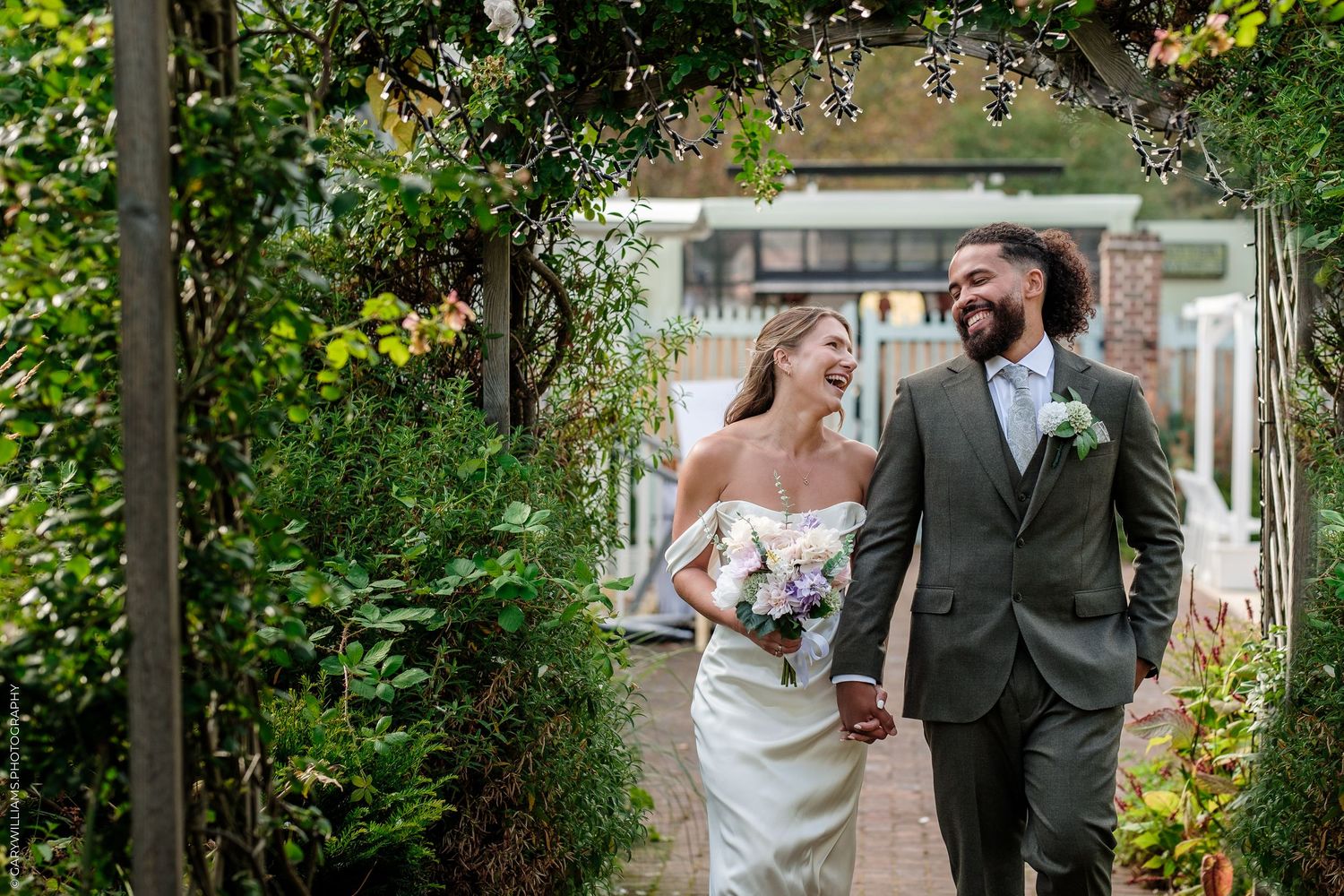 A joyful bride and groom walk hand-in-hand through a lush garden archway, laughing together on their wedding day.