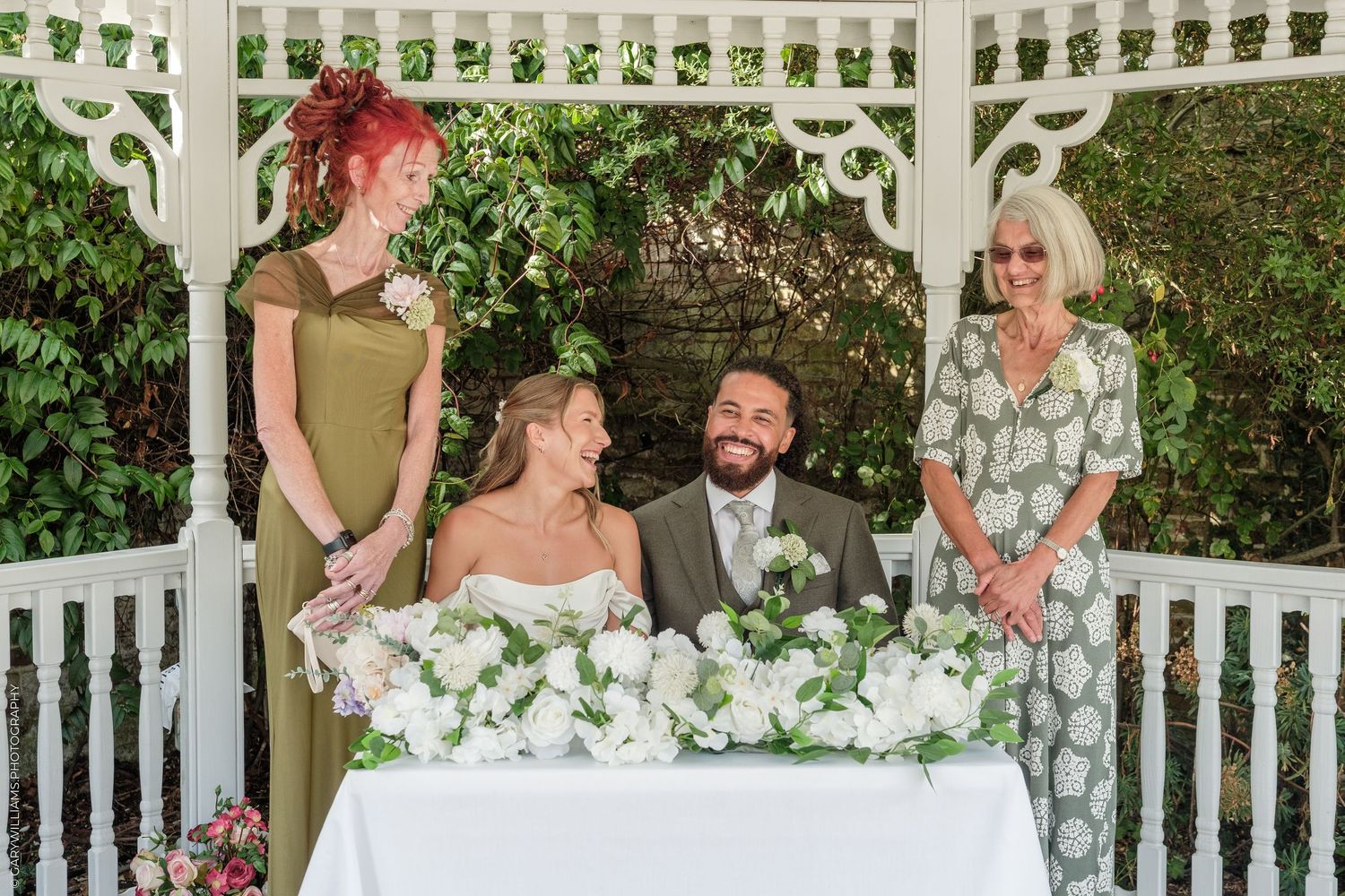 Couple signing wedding register in gazebo with bridesmaid and officiant, surrounded by white floral arrangements.