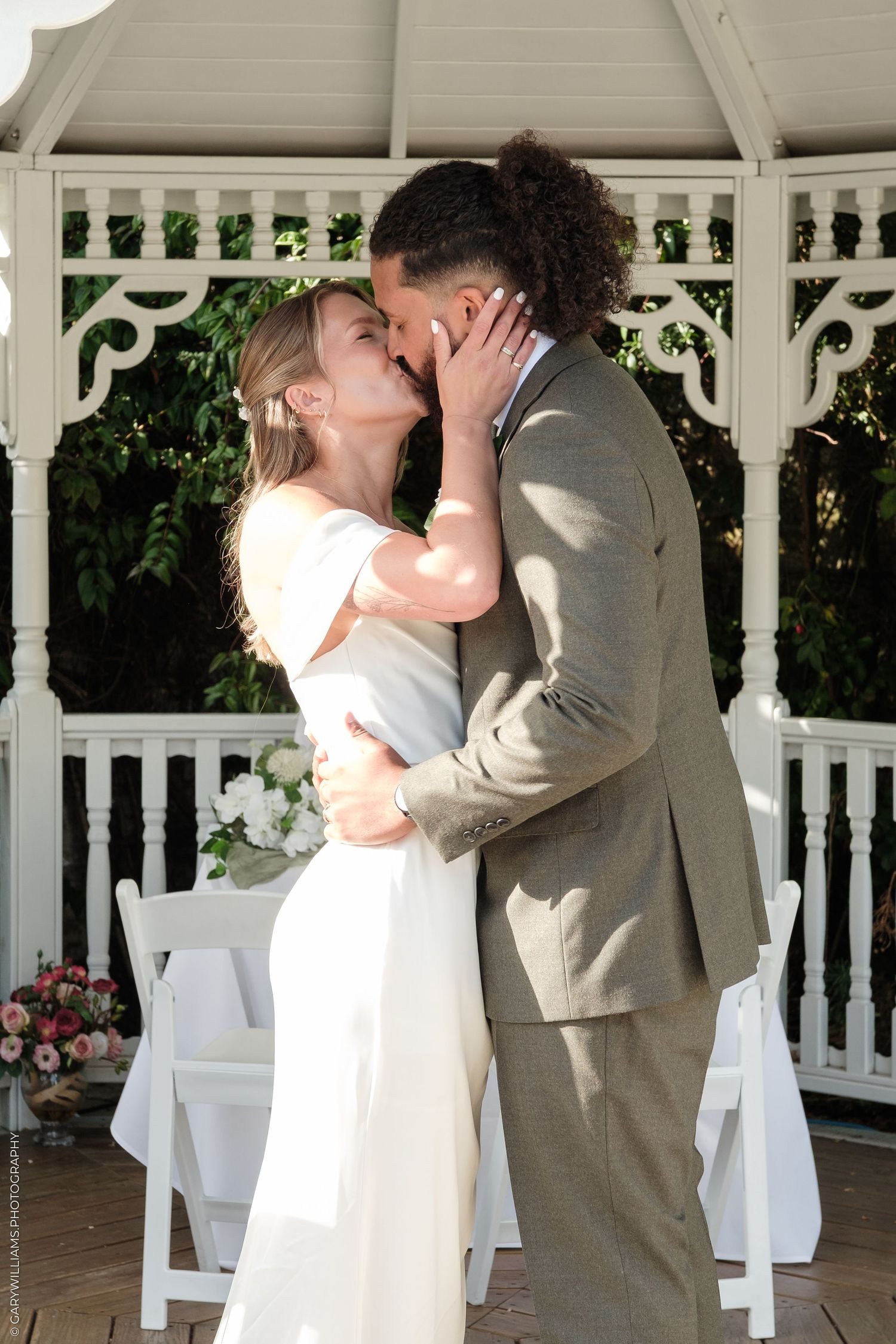 Interracial couple sharing first kiss at outdoor gazebo wedding ceremony, bride in white gown, groom in gray suit.