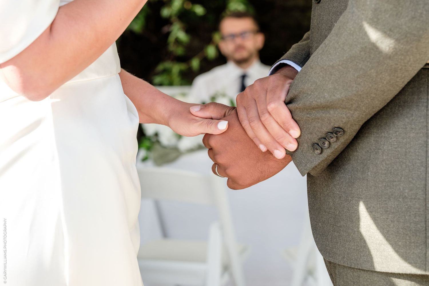 Bride and groom holding hands during wedding ceremony with officiant in background.