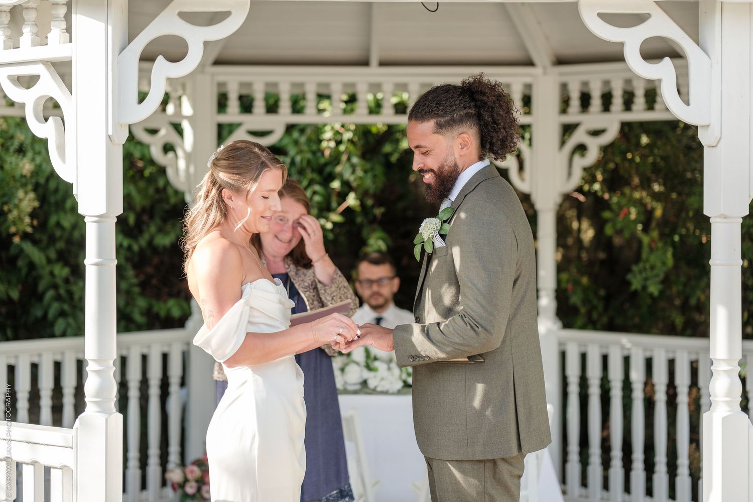 Bride and groom exchanging vows under a white gazebo, with the bride smiling and wiping tears during the wedding ceremony.