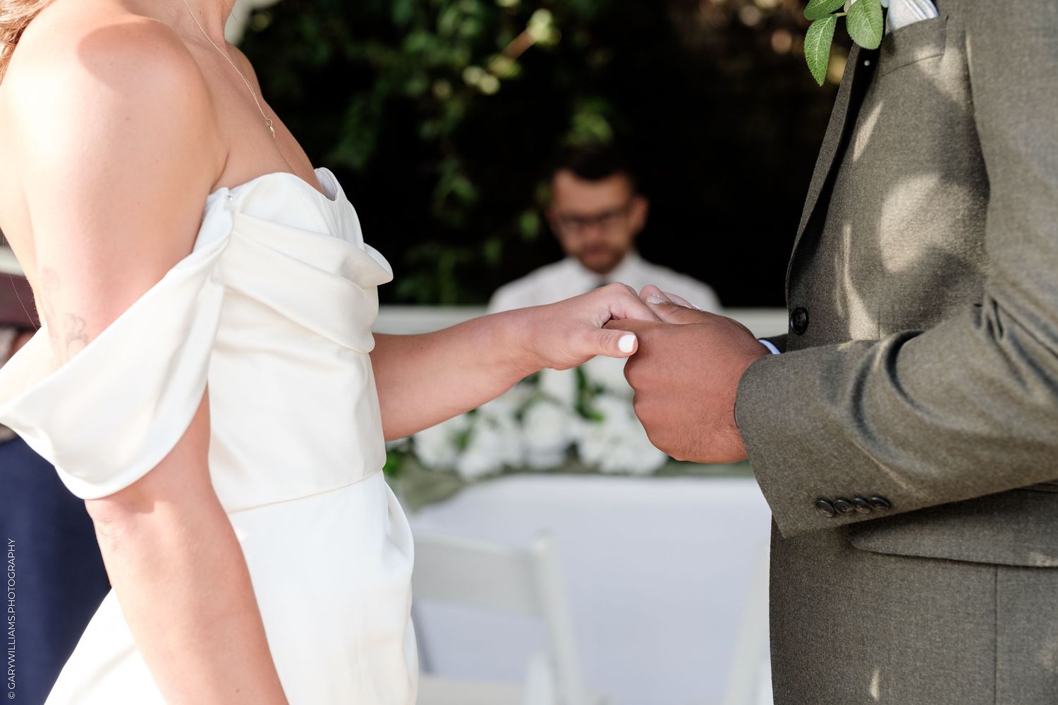 Bride placing a wedding ring on groom's finger during outdoor ceremony, officiant visible in background.