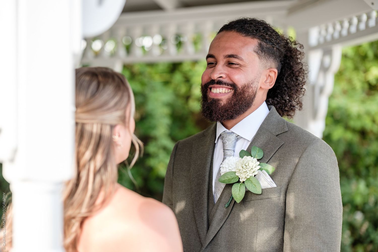 Groom smiling joyfully at bride during outdoor wedding ceremony, wearing tan suit with white rose boutonniere.