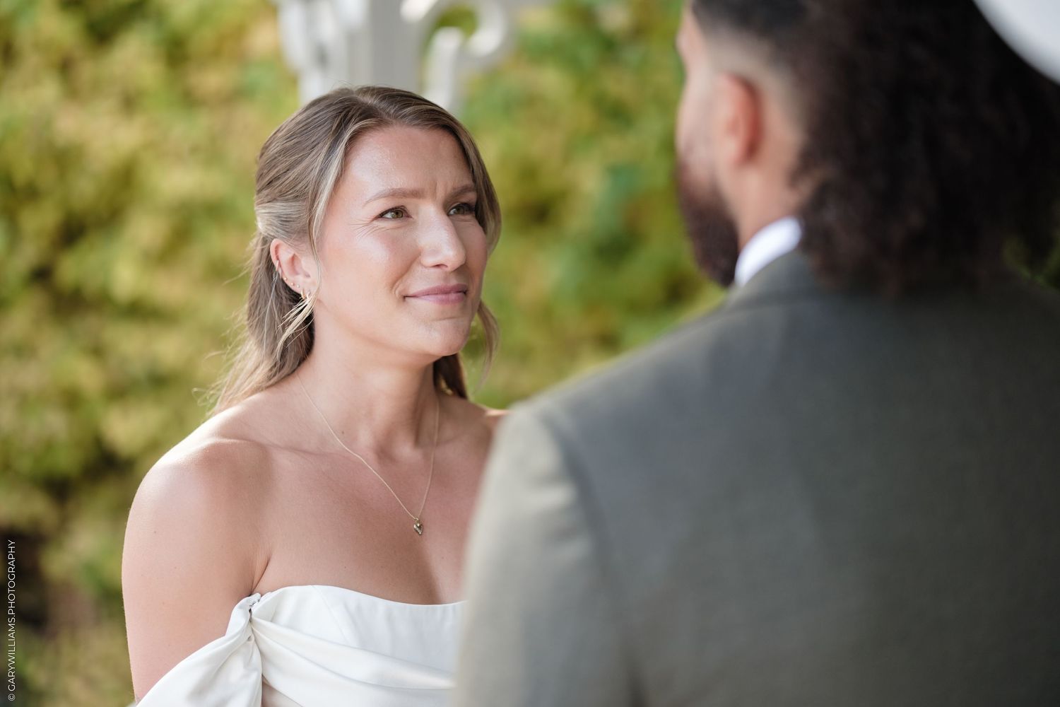Bride in white strapless gown smiling emotionally at groom during outdoor wedding ceremony.