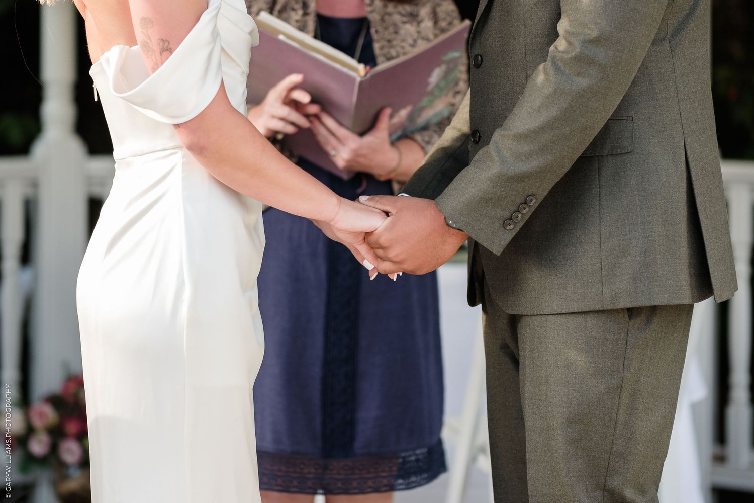 Bride and groom holding hands during outdoor wedding ceremony while officiant reads from book.