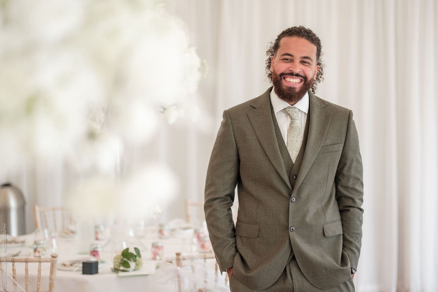 Smiling man with beard wearing olive green suit at elegant wedding reception venue with white floral decor.
