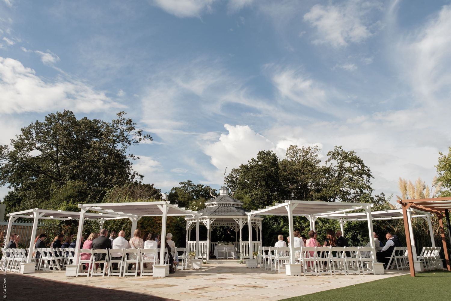 Outdoor wedding ceremony under white gazebo and pergolas on a sunny day with blue skies and lush green trees.