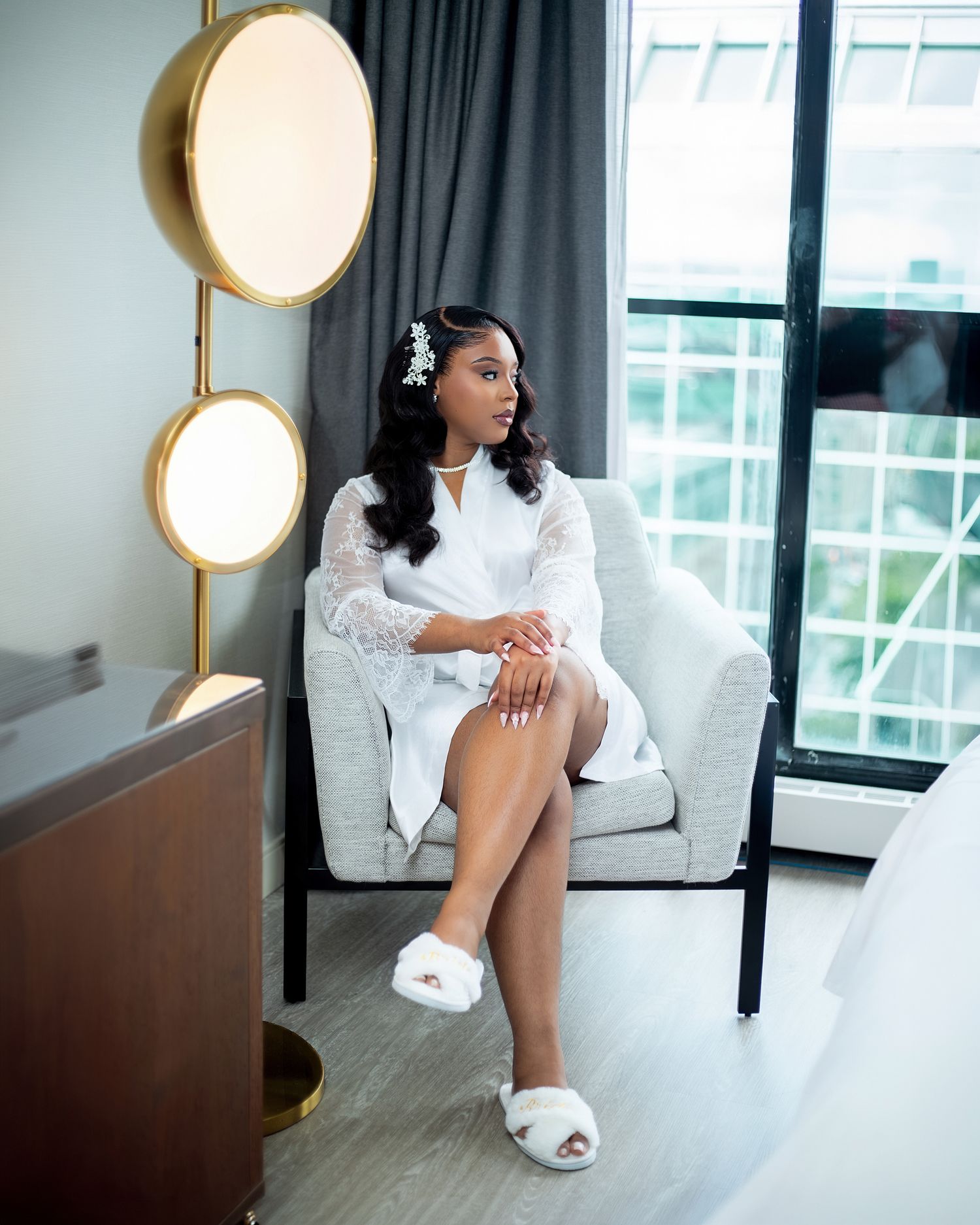 Bride in white lace robe and fluffy slippers sits elegantly in a modern hotel room chair before her wedding.