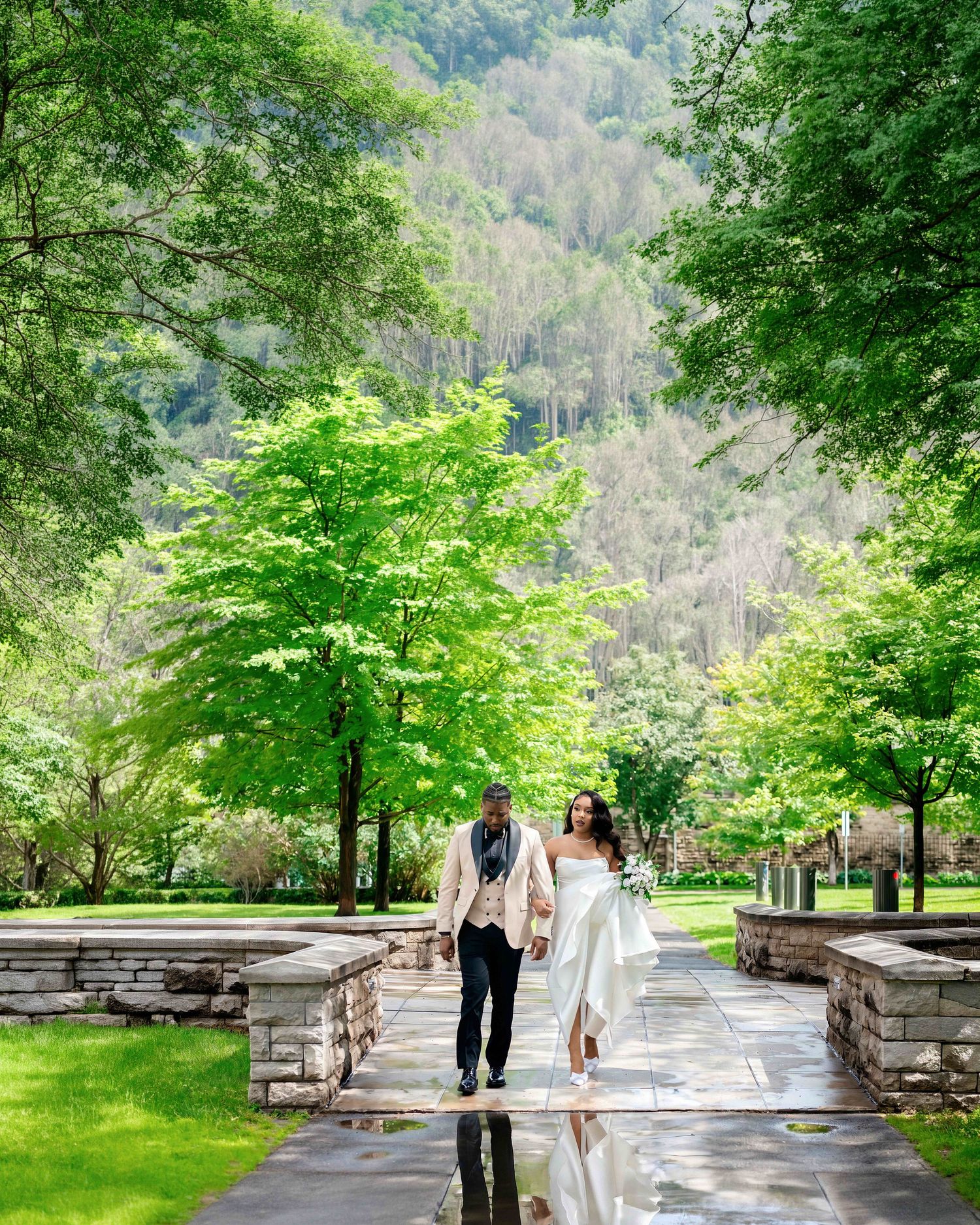 Bride and groom walk hand-in-hand along a stone path surrounded by lush green trees with a mountain backdrop.
