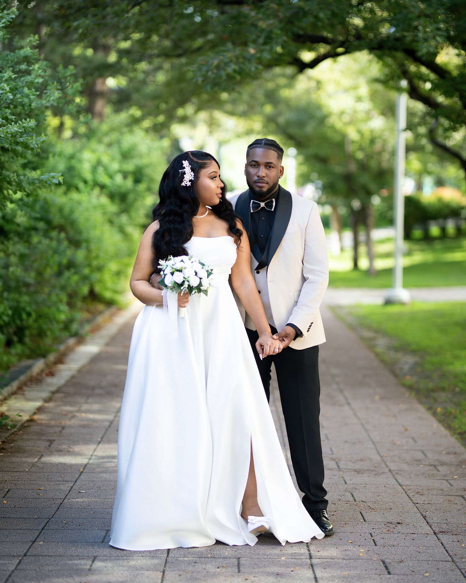 A bride in a white gown holds a bouquet while a groom in a white tuxedo jacket holds her hand on a tree-lined path.