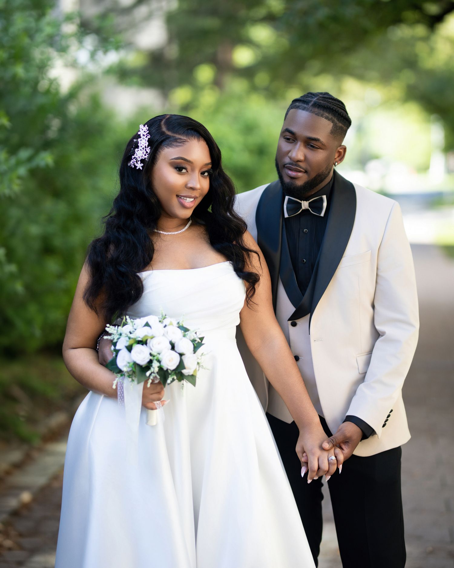A bride in a white strapless gown holds a bouquet beside a groom in a cream tuxedo with black bow tie outdoors.