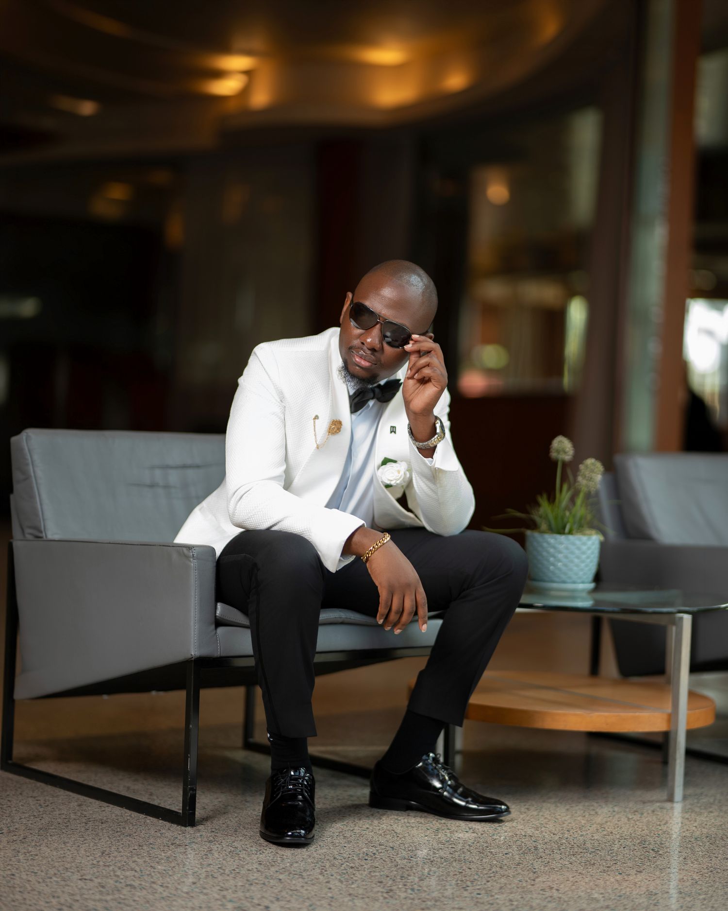Stylish man in white blazer and bow tie sitting on gray sofa, adjusting sunglasses in modern lobby.
