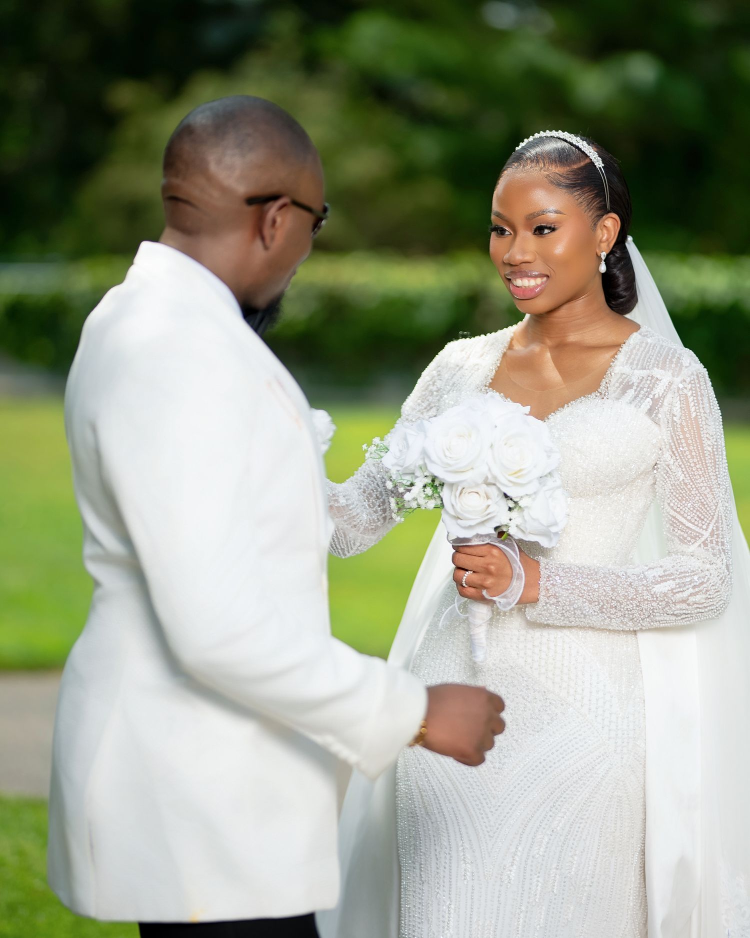 A bride in a beaded white gown holds a bouquet while sharing a tender moment with her groom in a white suit outdoors.