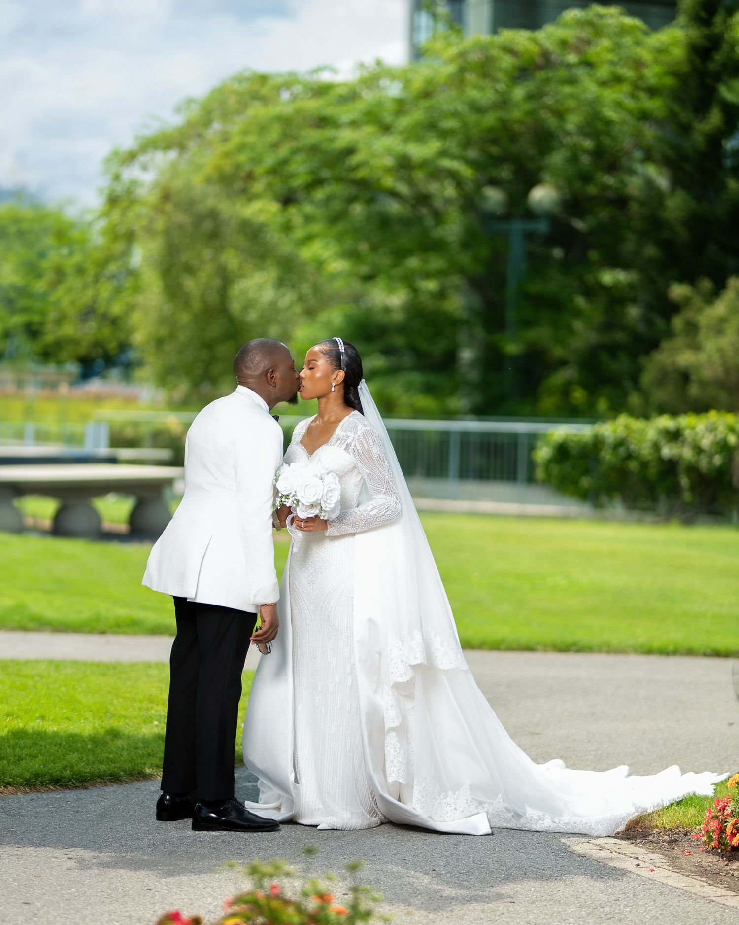 Bride and groom sharing a kiss in a lush garden, wearing white wedding attire with a flowing bridal train.