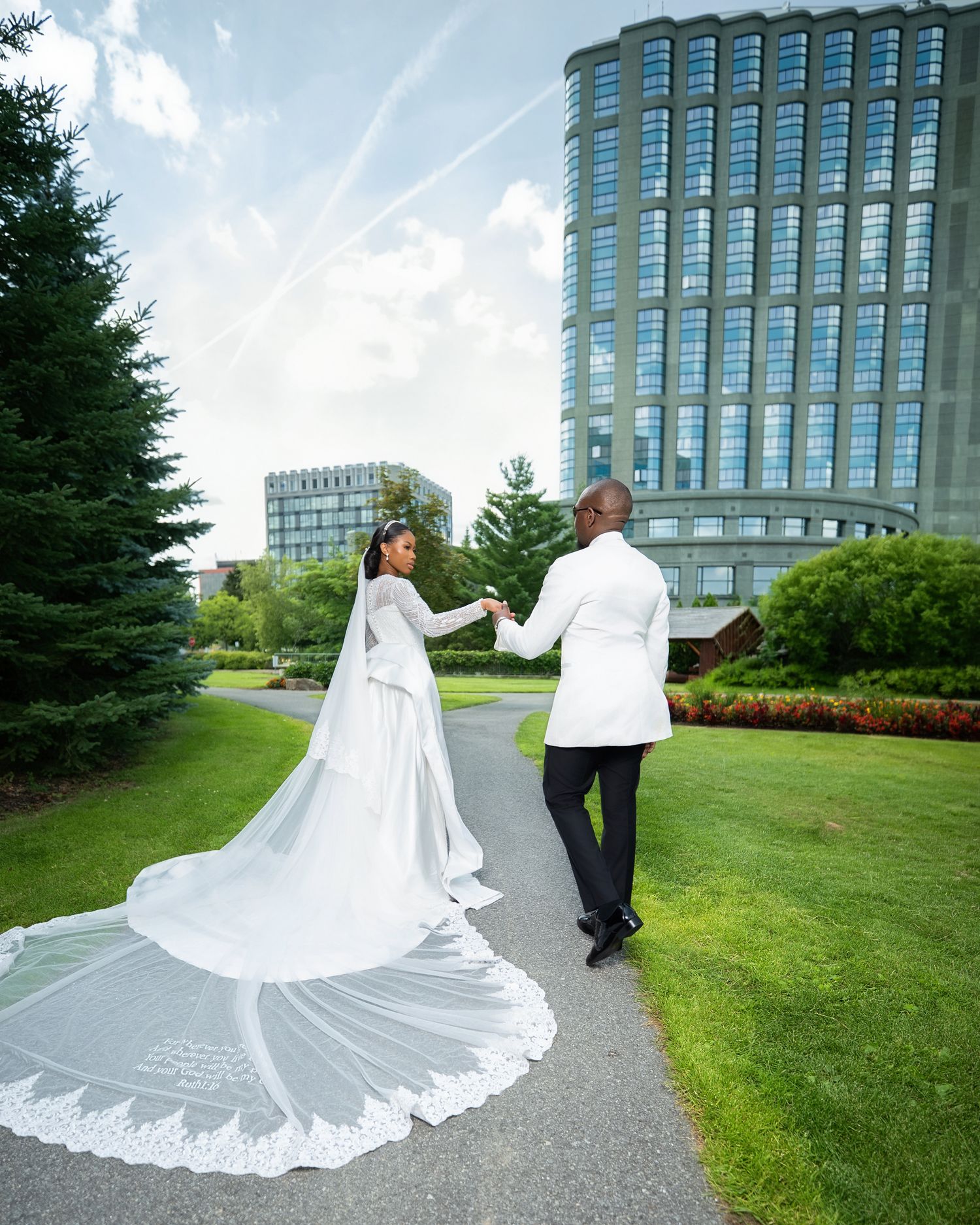 Bride and groom walking hand-in-hand on a garden path, her long lace cathedral veil trailing behind, with a glass tower building backdrop.