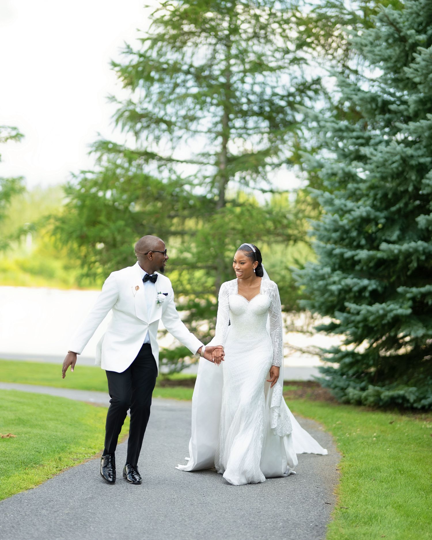 A bride and groom in white wedding attire hold hands while walking on a garden path surrounded by lush green trees.