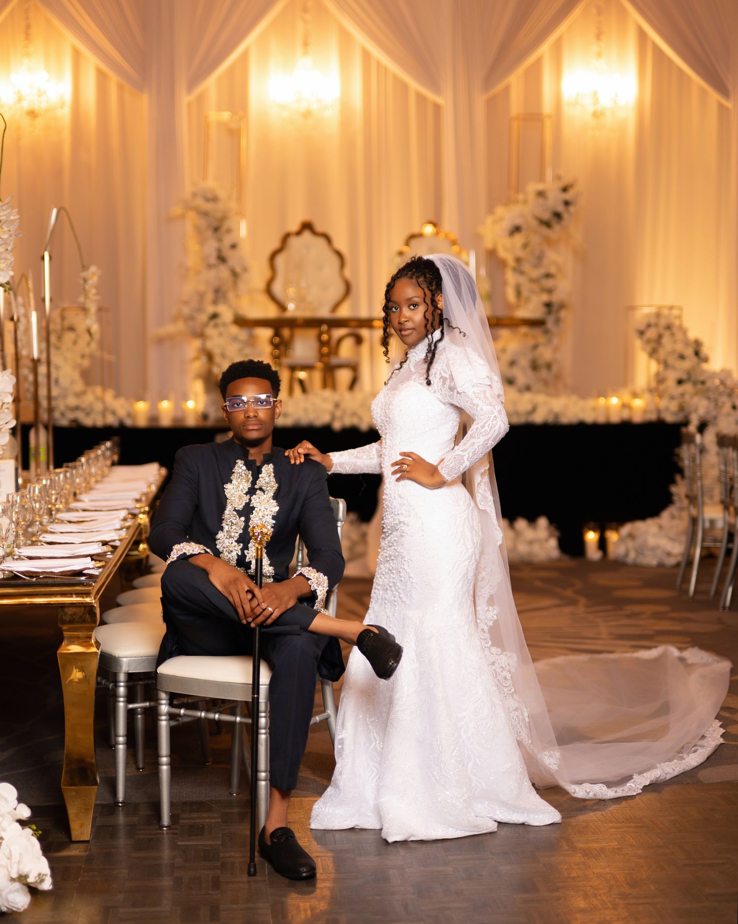 Elegant wedding couple posing in a luxurious white-draped venue, groom in black suit, bride in lace gown with veil.