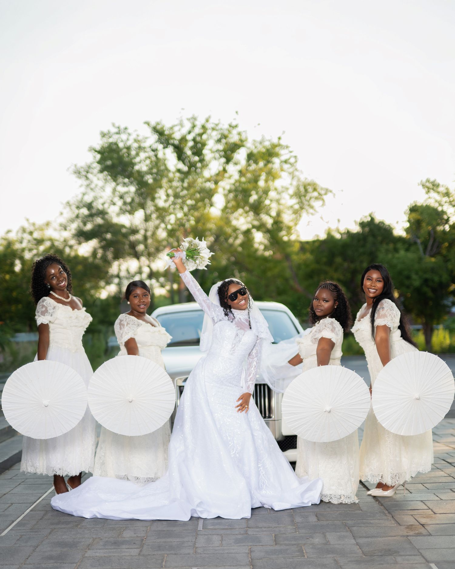 Bride in white gown raises bouquet with four bridesmaids holding white parasols beside a white limo outdoors.