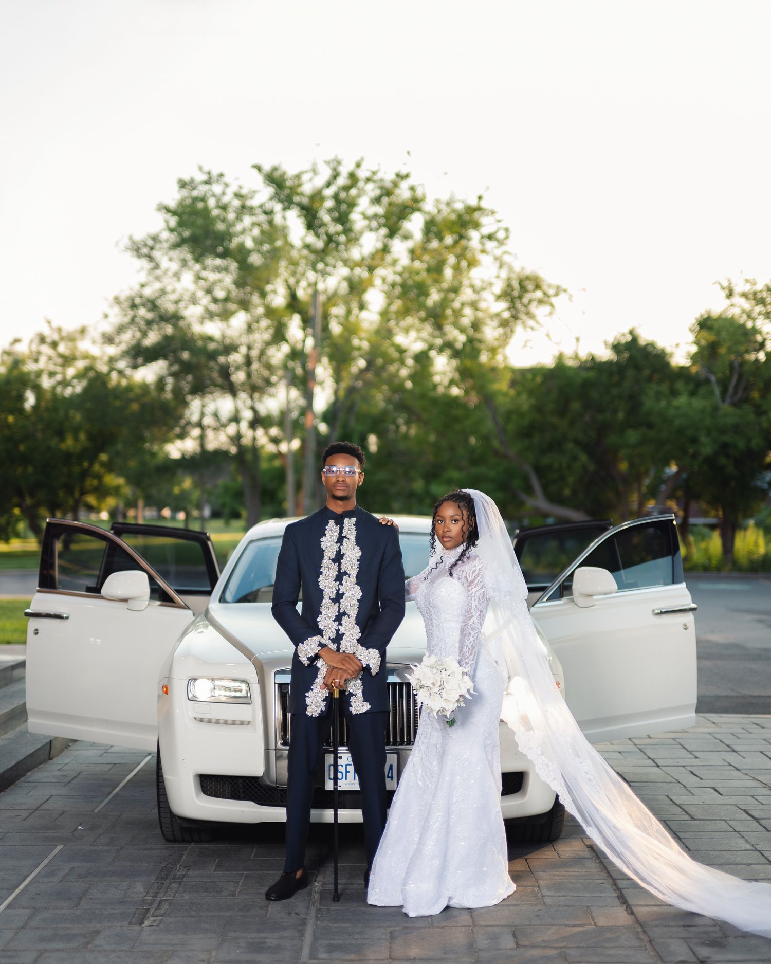 Elegant bride and groom in wedding attire pose in front of a white Rolls-Royce on their wedding day.