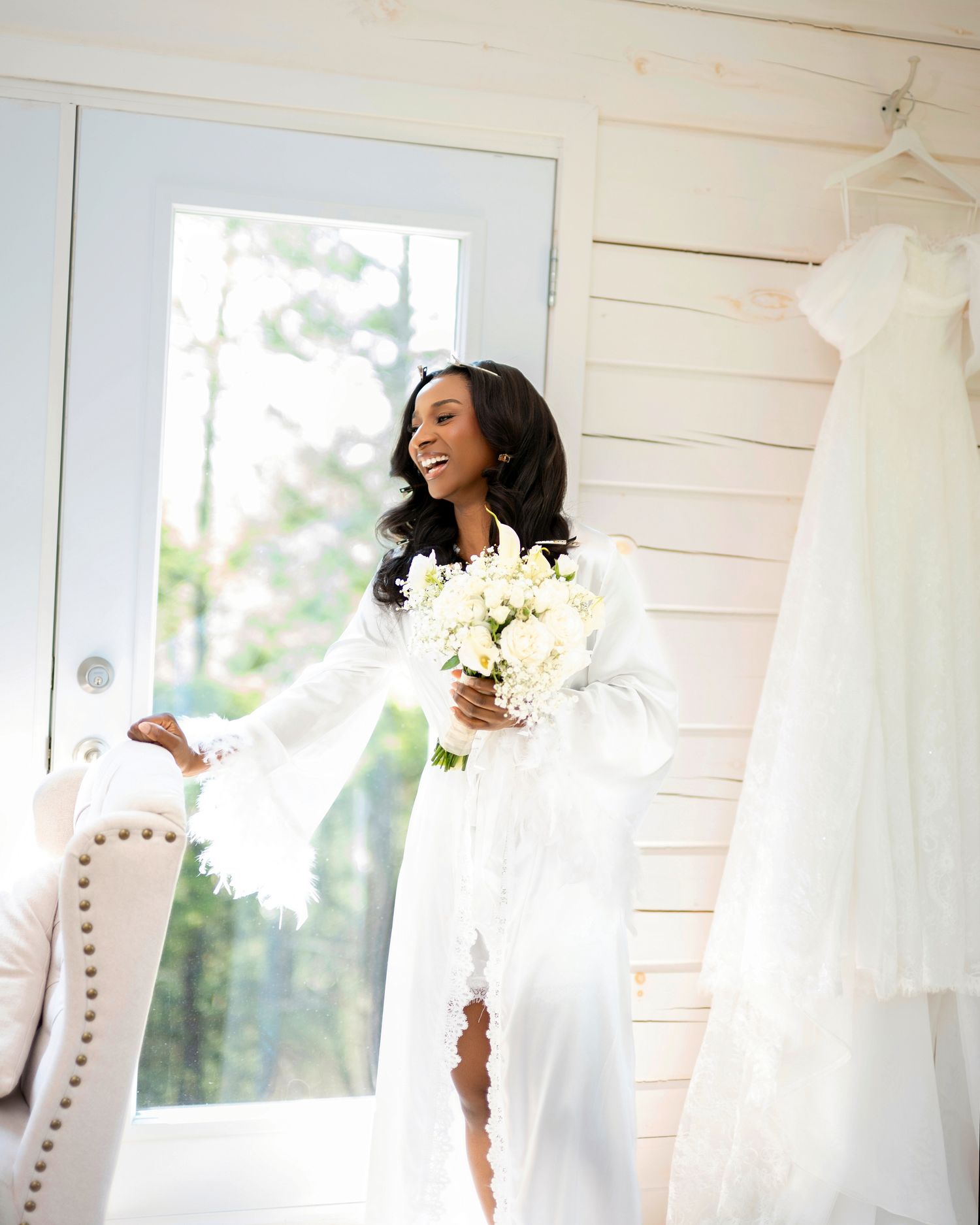 A radiant Black bride in a white lace robe holds a bouquet near her wedding gown hanging in a bright, elegant dressing room.