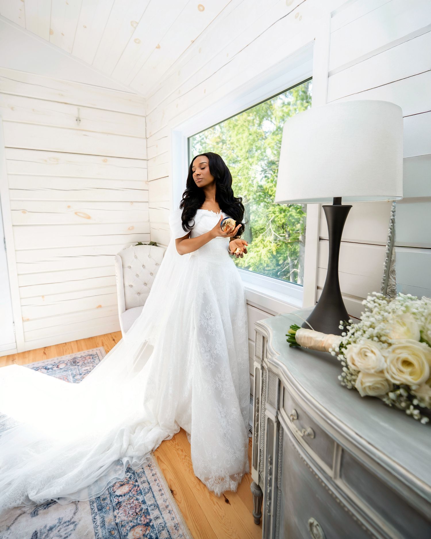 Bride in elegant white off-shoulder ball gown with long train stands by window in rustic white shiplap bridal suite.