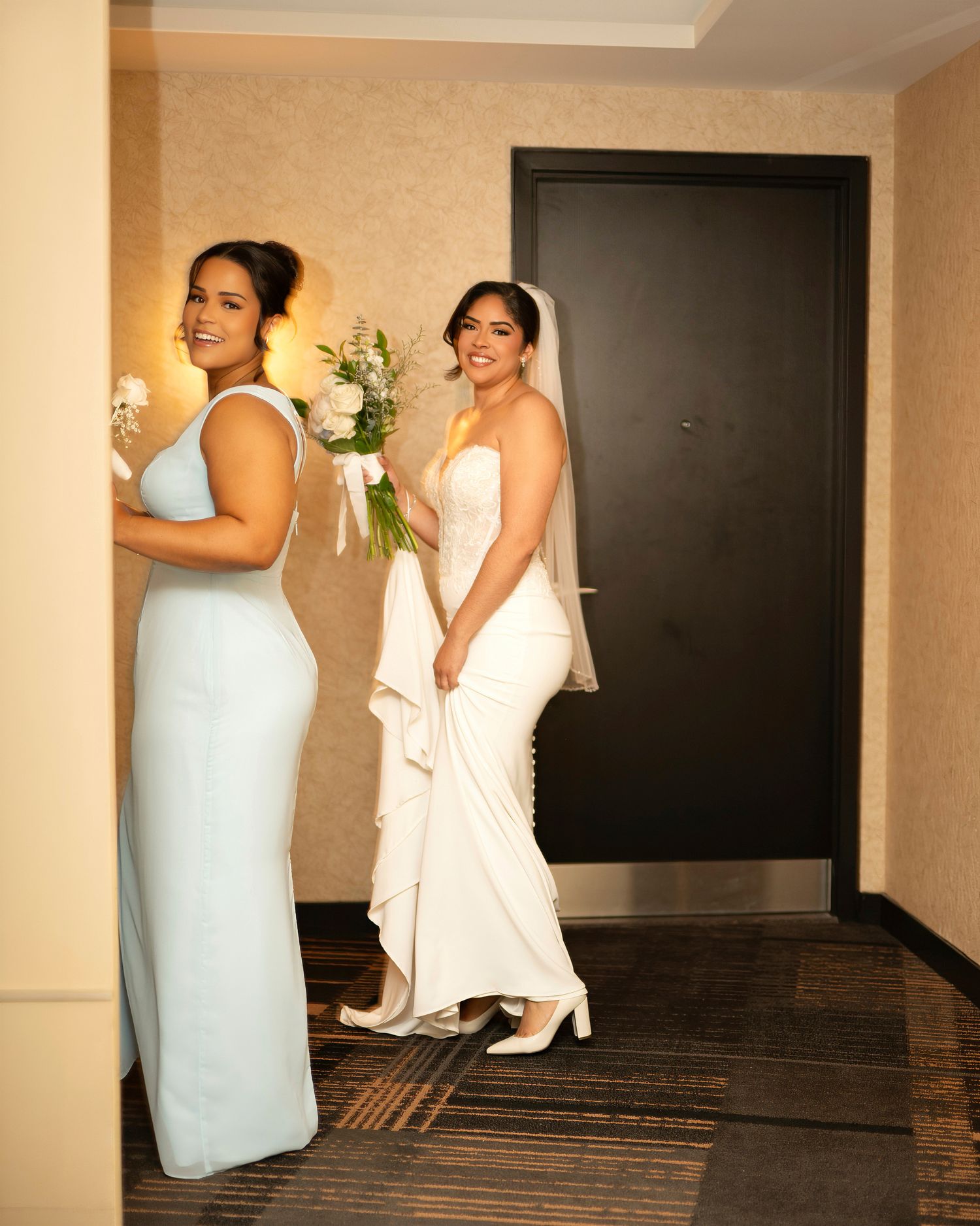 Bride in white gown holding bouquet with bridesmaid in light blue dress standing in hotel hallway.