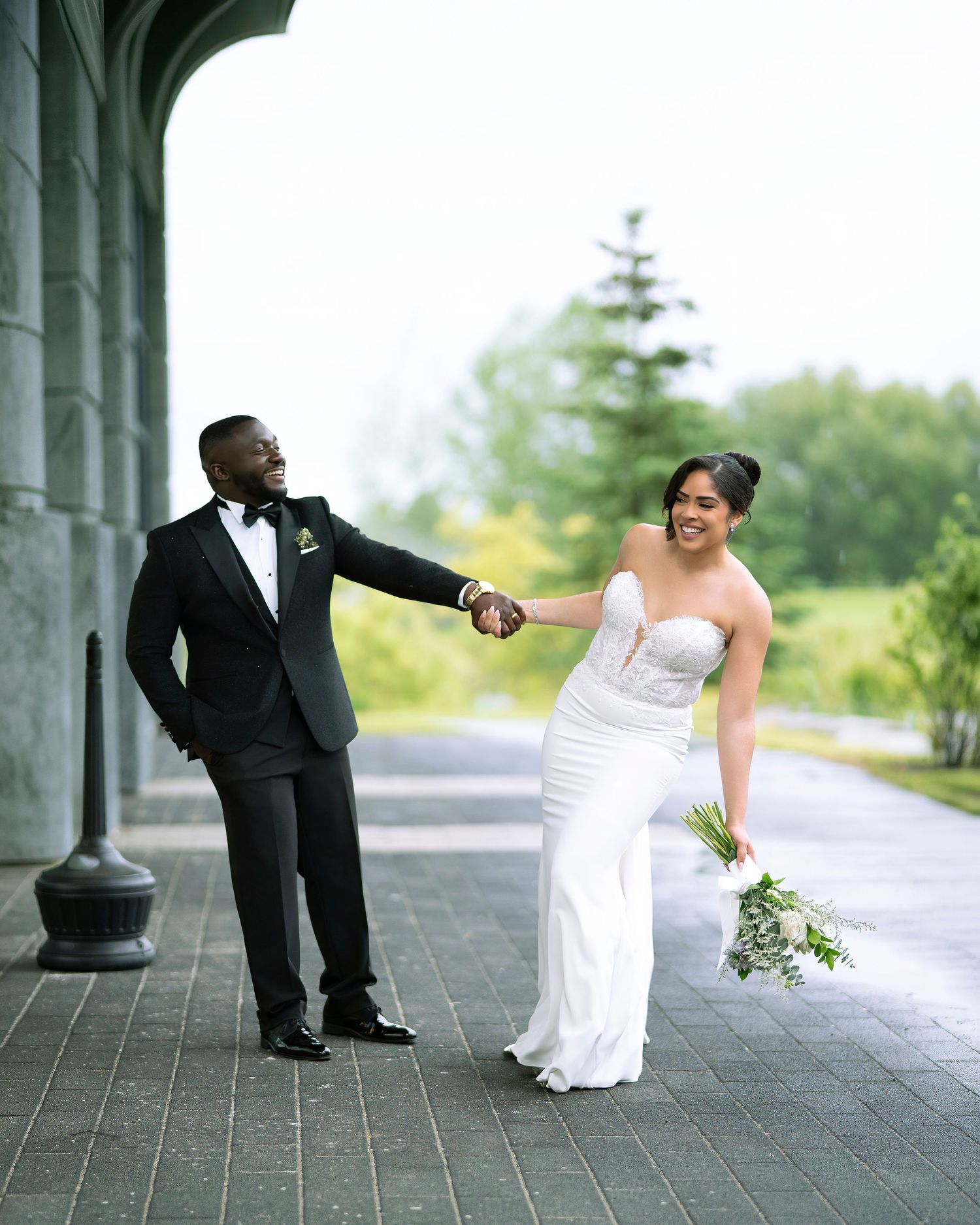 A groom in a black tuxedo holds his bride's hand as she walks in a white gown holding greenery bouquet outdoors.