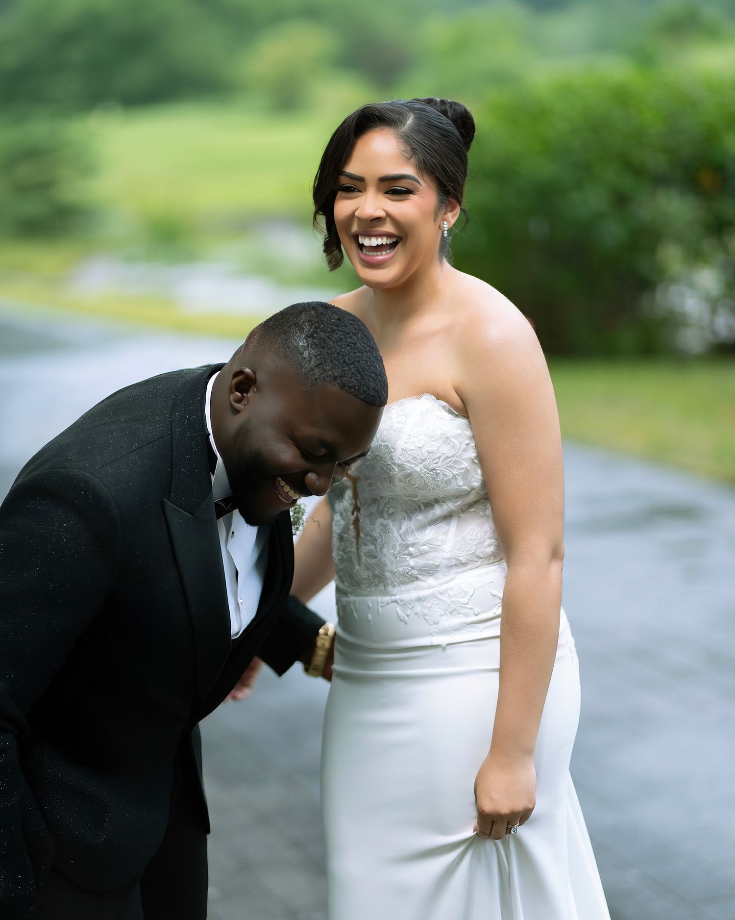 Joyful bride in strapless white gown laughs with groom in black tuxedo on a scenic outdoor path.