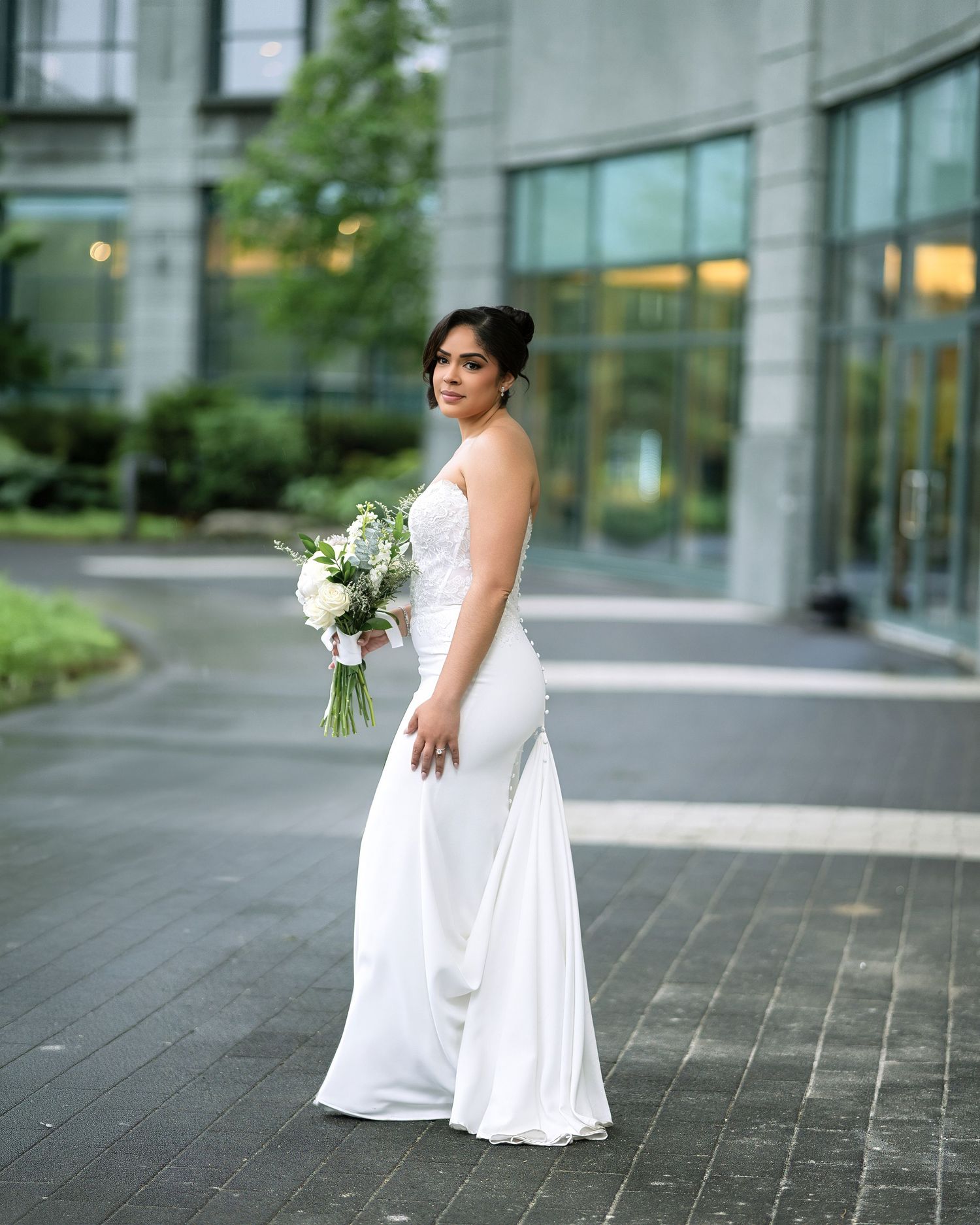 Bride in white gown holding bouquet of white flowers, posing outdoors near modern glass building.