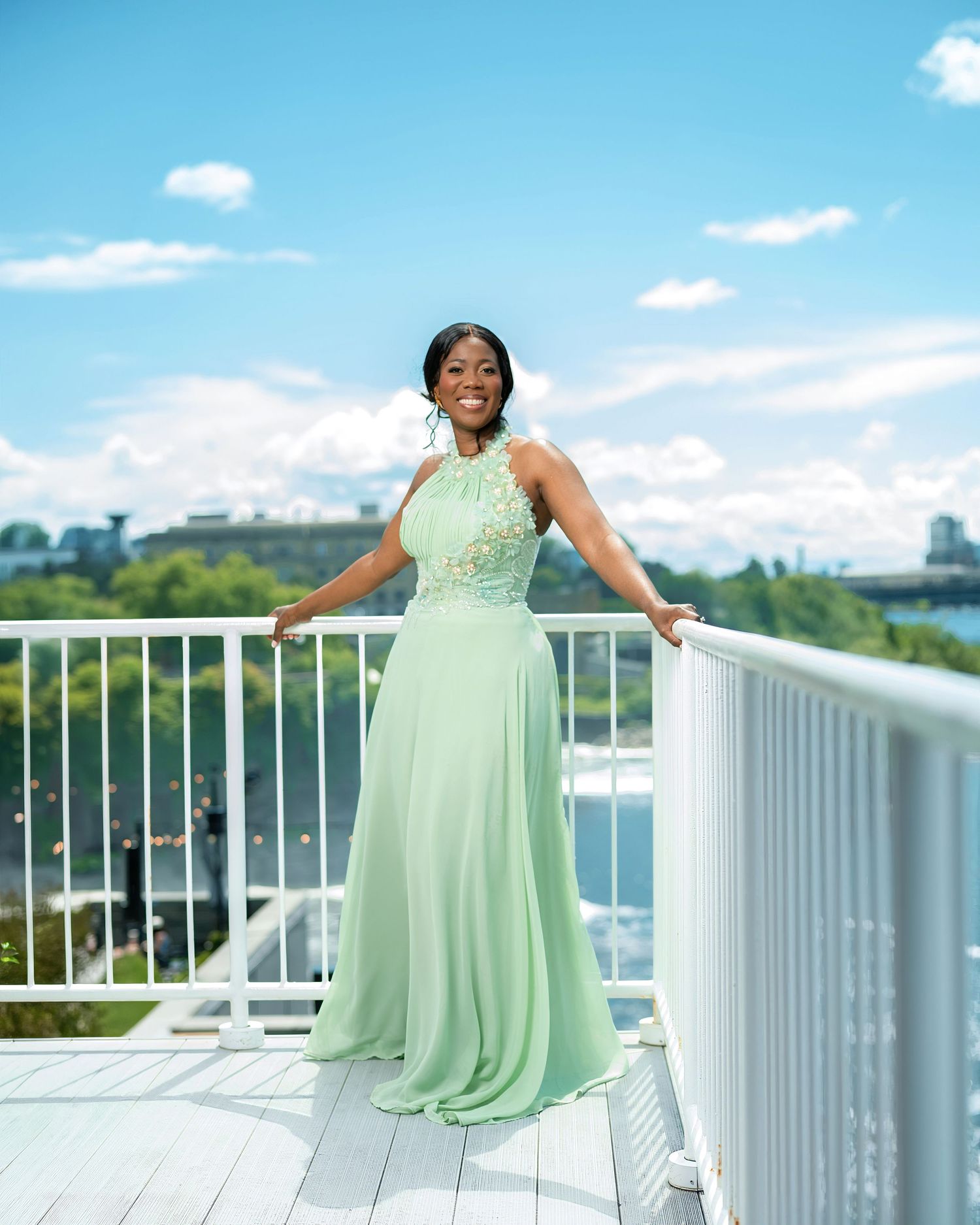 Woman in mint green halter gown smiling on white balcony overlooking waterfront on sunny day.