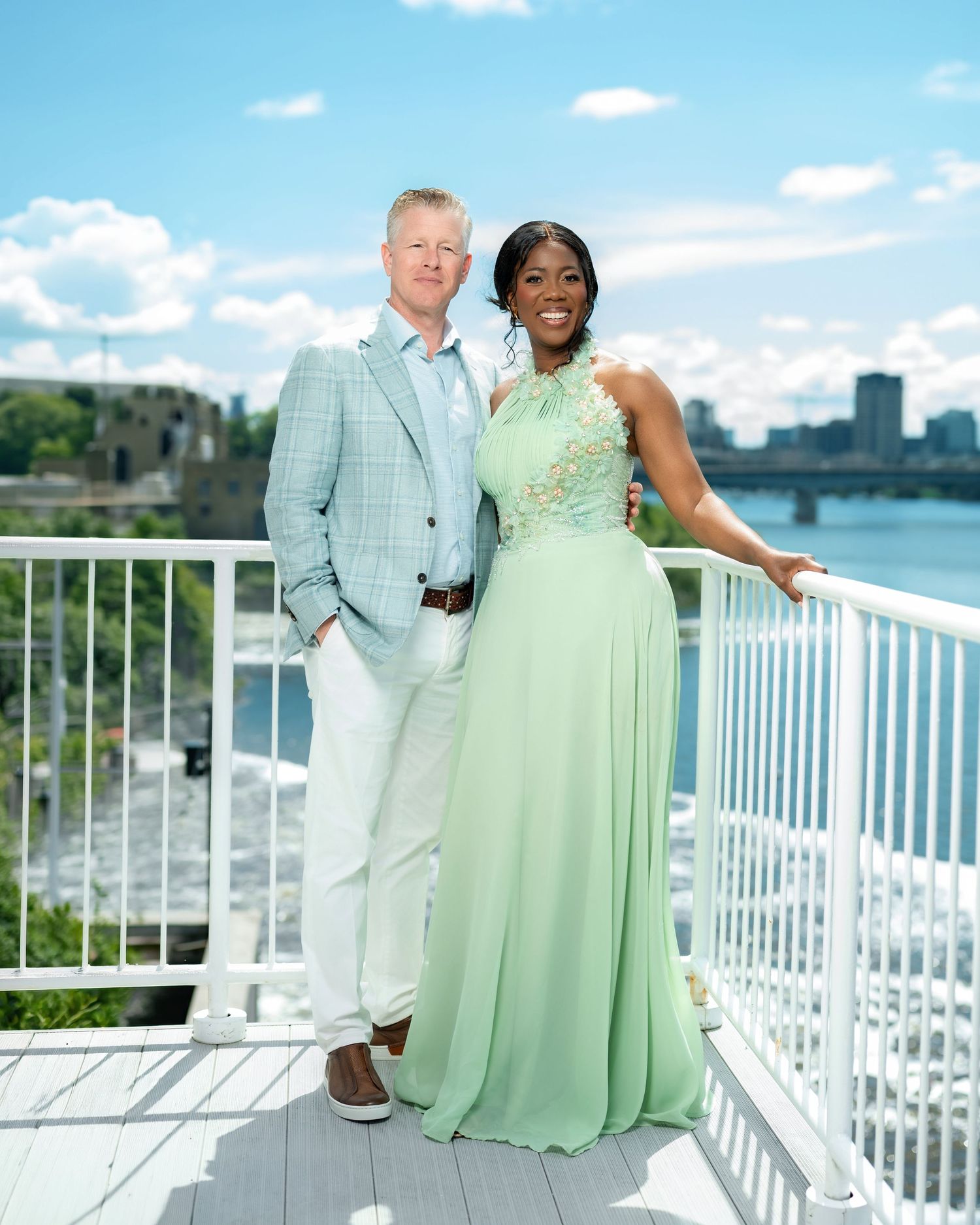 Elegant couple posing on a waterfront deck, man in plaid blazer, woman in mint green gown, city skyline behind.