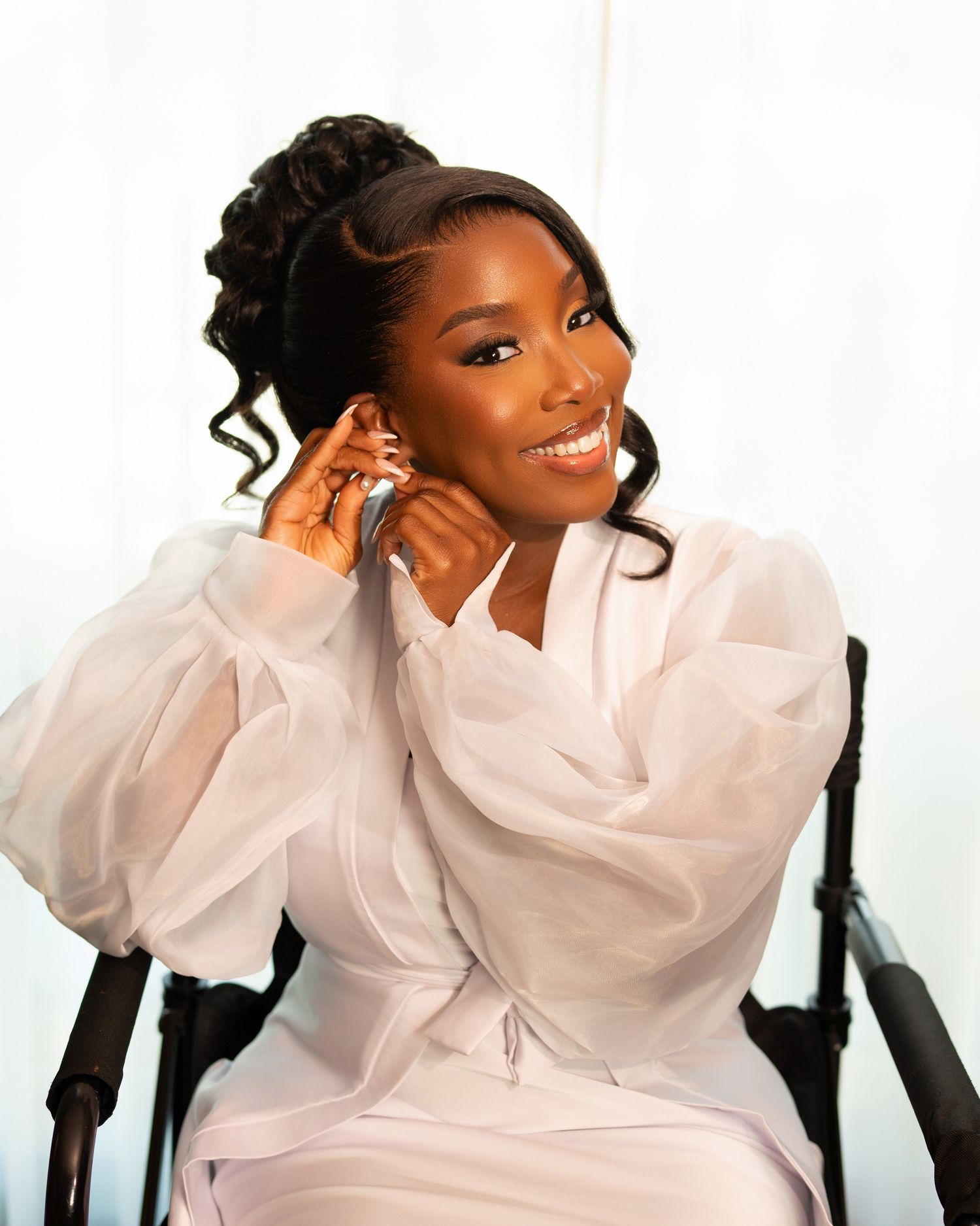 A smiling woman in a white sheer puff-sleeve dress poses gracefully with hands near her face against a white background.