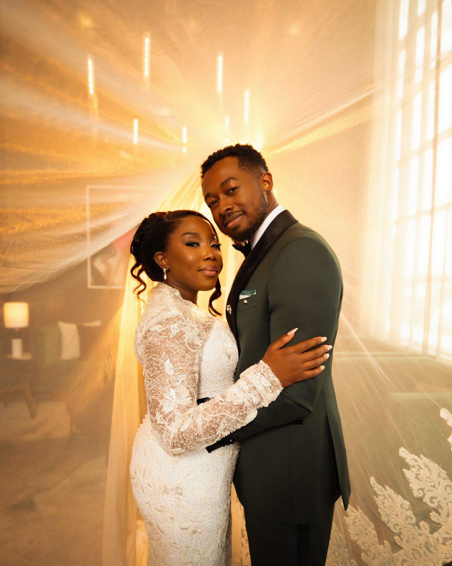 A bride in a lace gown and groom in a dark suit embrace elegantly in a warmly lit wedding venue.