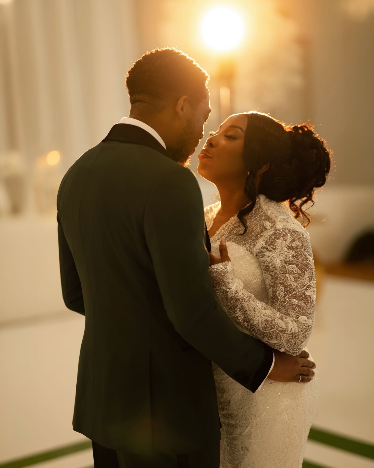 A bride and groom share a romantic embrace in golden backlight, she in lace gown, he in black suit.