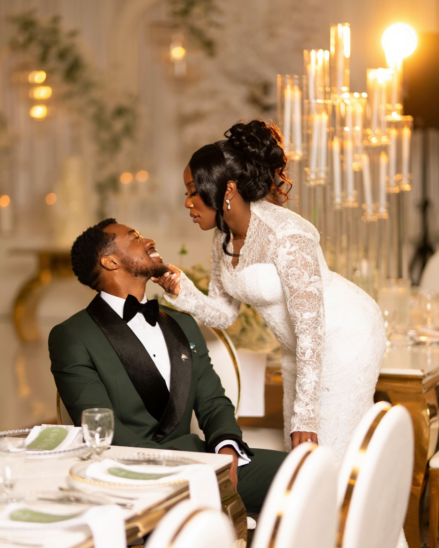Bride in lace gown tenderly touches groom's face as they share an intimate moment at elegant wedding reception.