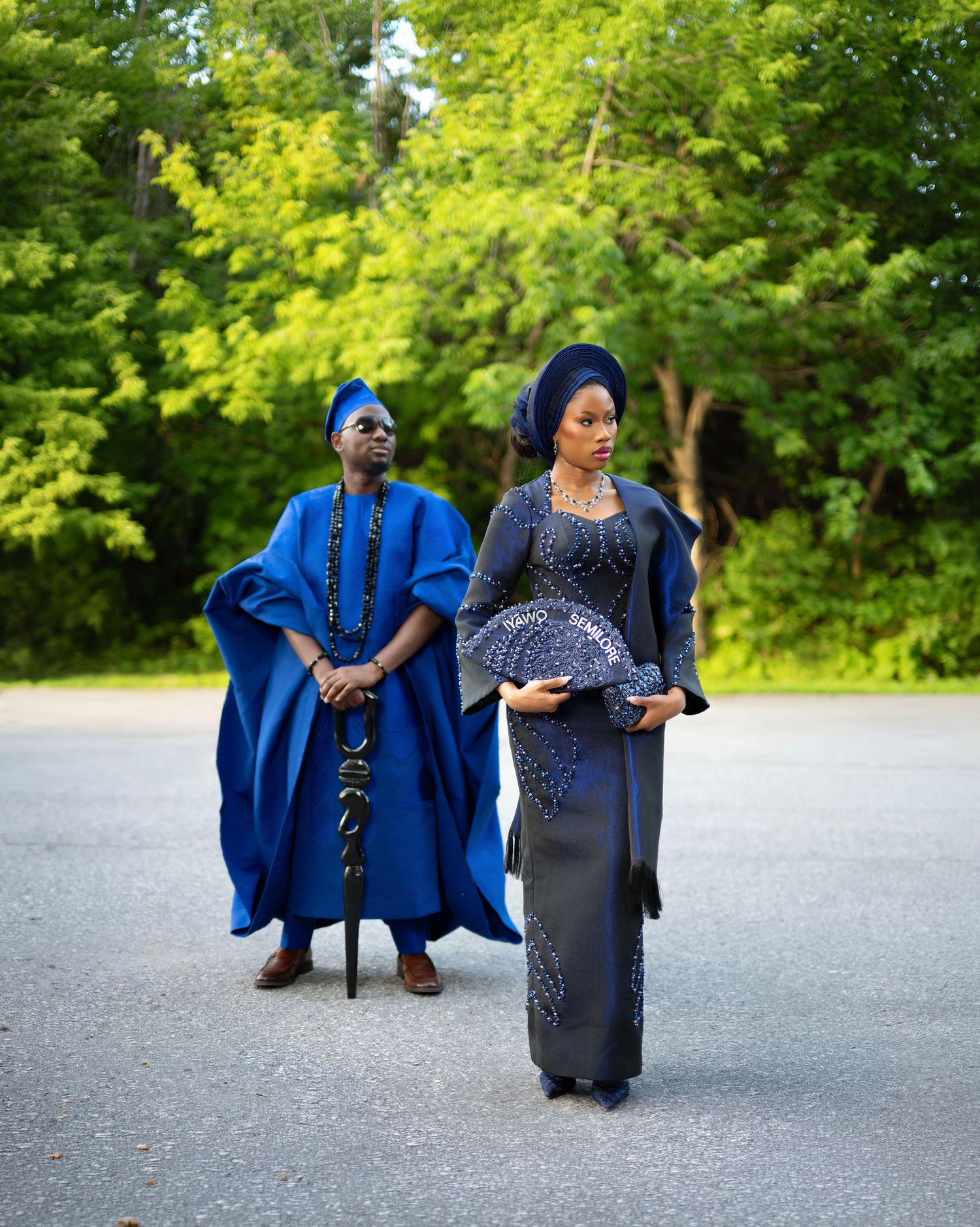 Couple in traditional Nigerian Yoruba attire, man in blue agbada with cap, woman in elegant navy embellished gown outdoors.